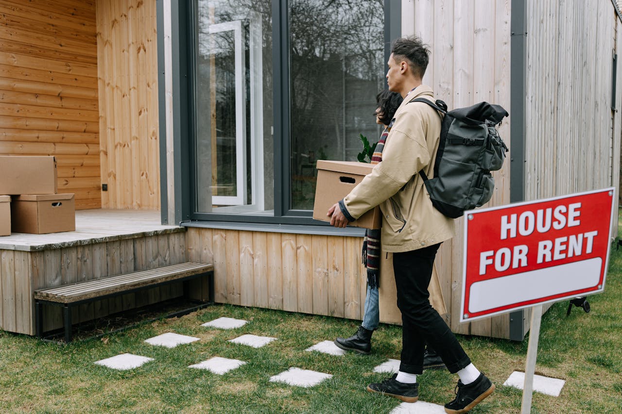 hero-img-01 Young couple moving into a new rental home with boxes. Outdoor scene with a rental sign.